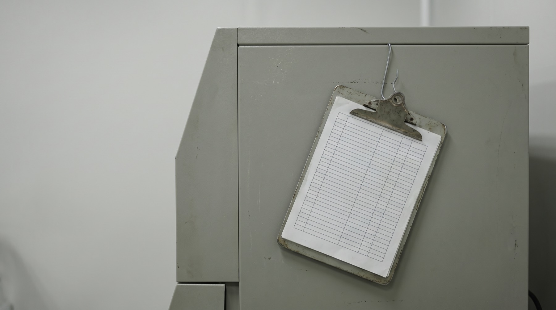 A clipboard holding a ruled log sheet, hanging beside a plain benchtop instrument